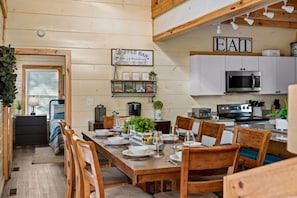 A cozy, well-lit dining area with a wooden table set for four, surrounded by chairs. A sign above the table reads "Coffee Bar" and lists beverages. The room features a kitchen with white cabinets and a microwave.