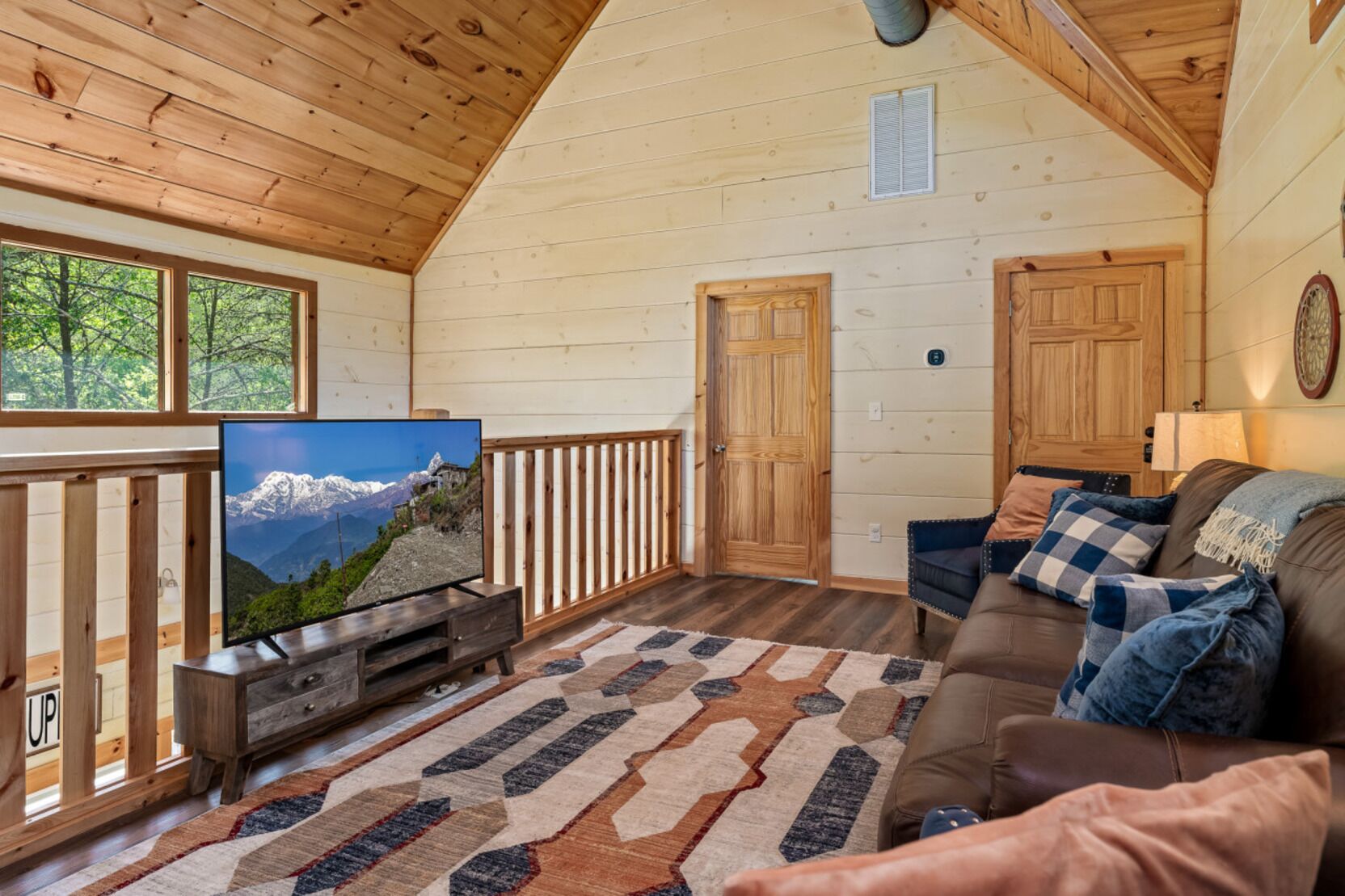 A cozy cabin interior with a wooden ceiling, a television displaying a mountain view, a brown leather sofa, and a patterned rug.