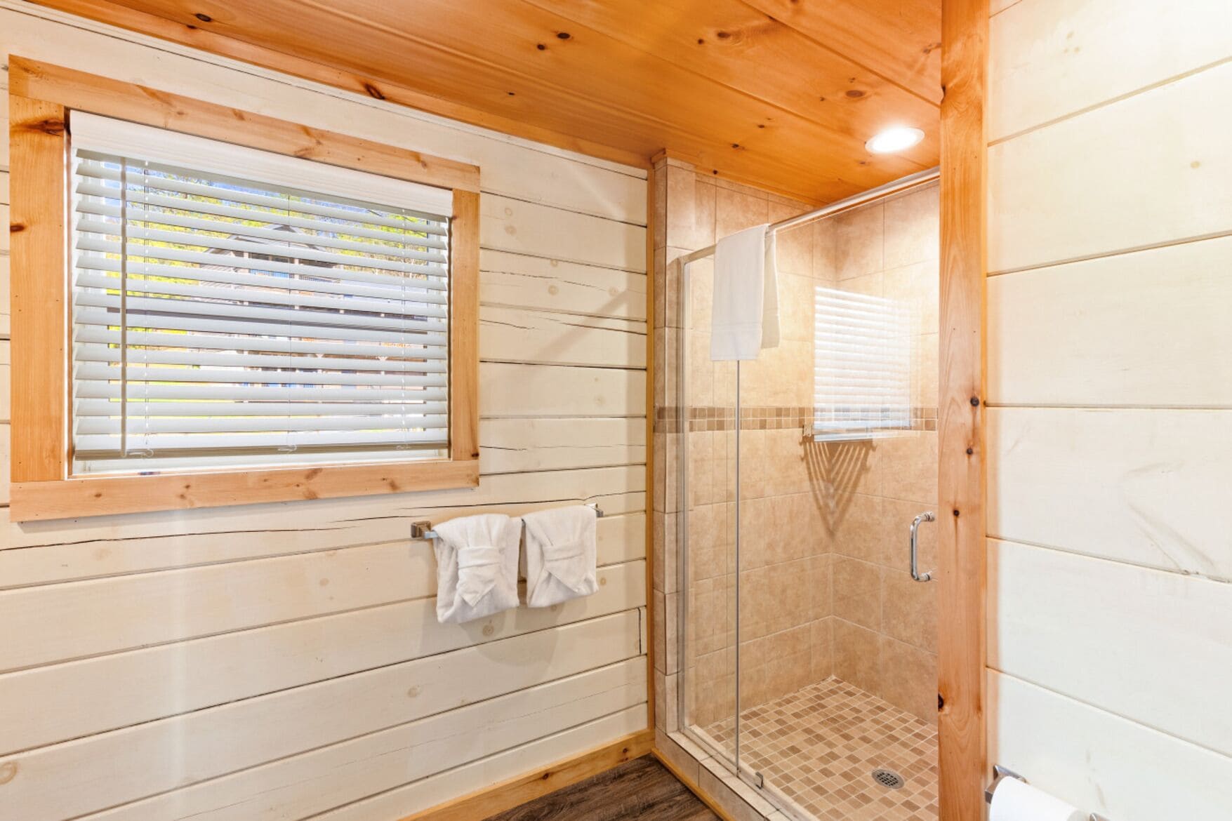 Une salle de bain aux murs lambrissés, dotée d’une douche en verre, d’une fenêtre avec stores et de deux serviettes suspendues à un porte-serviettes.
