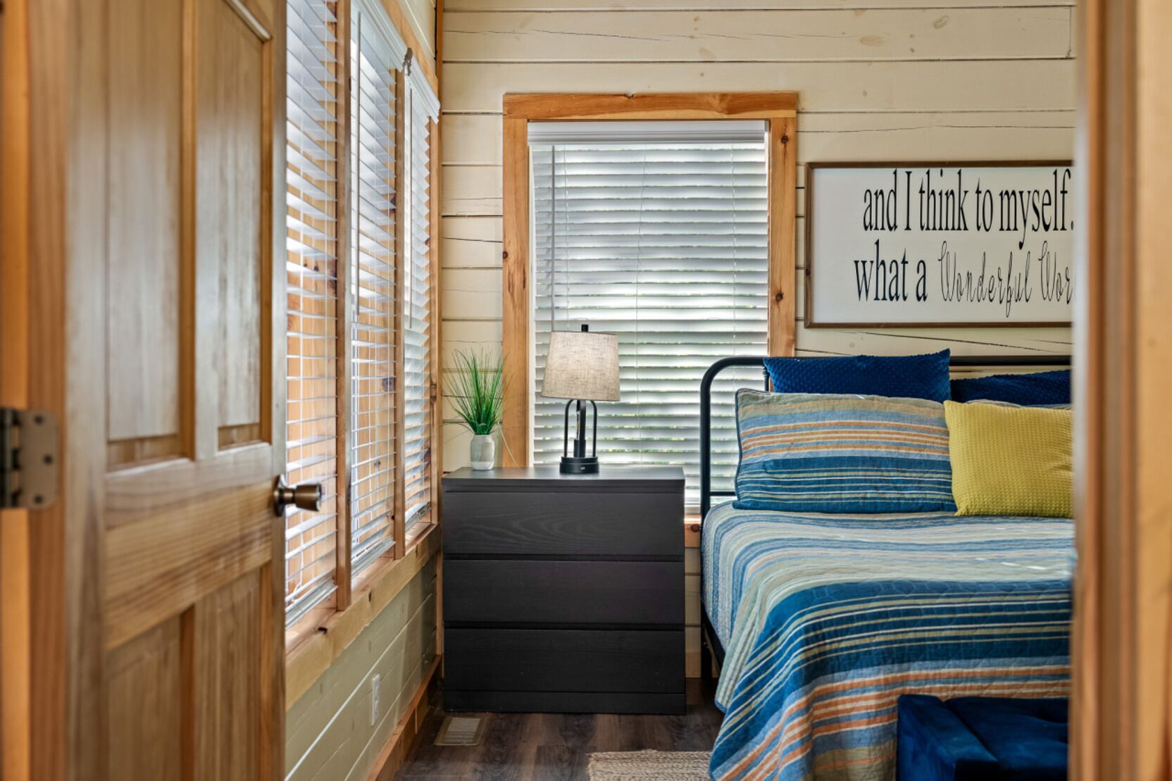 A bedroom with a bed, a nightstand, a lamp, a window with blinds, and a wall with a motivational quote.