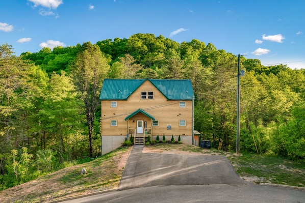 A two-story wooden house with a green roof, surrounded by dense forest.