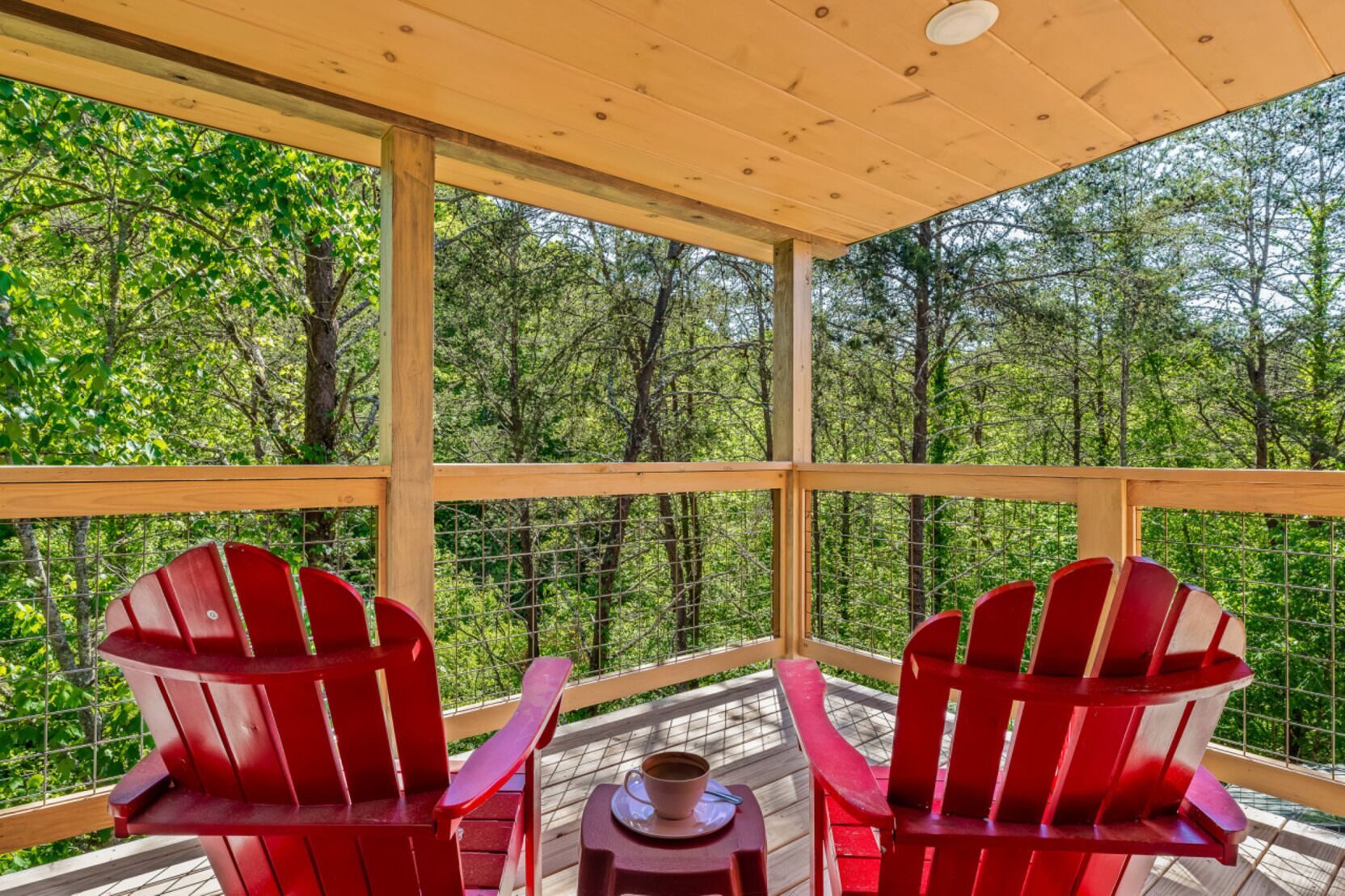 A covered outdoor seating area with red Adirondack chairs and a table with a cup and saucer.