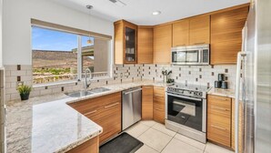 A modern kitchen with wooden cabinets, stainless steel appliances, a large window with a view, and a tiled backsplash. - Modern Resort Condo - Pools, Courts & Scenic Views (St. George)