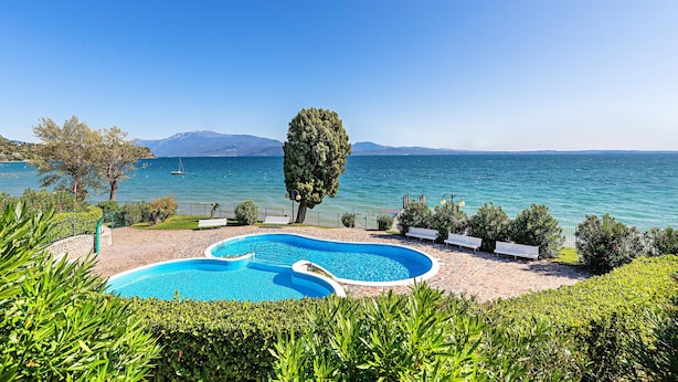 A swimming pool with a curved design, surrounded by a stone patio and benches, with a view of the sea and mountains in the background. - San Sivino 142 Apartment by Wonderful Italy (Manerba del Garda)
