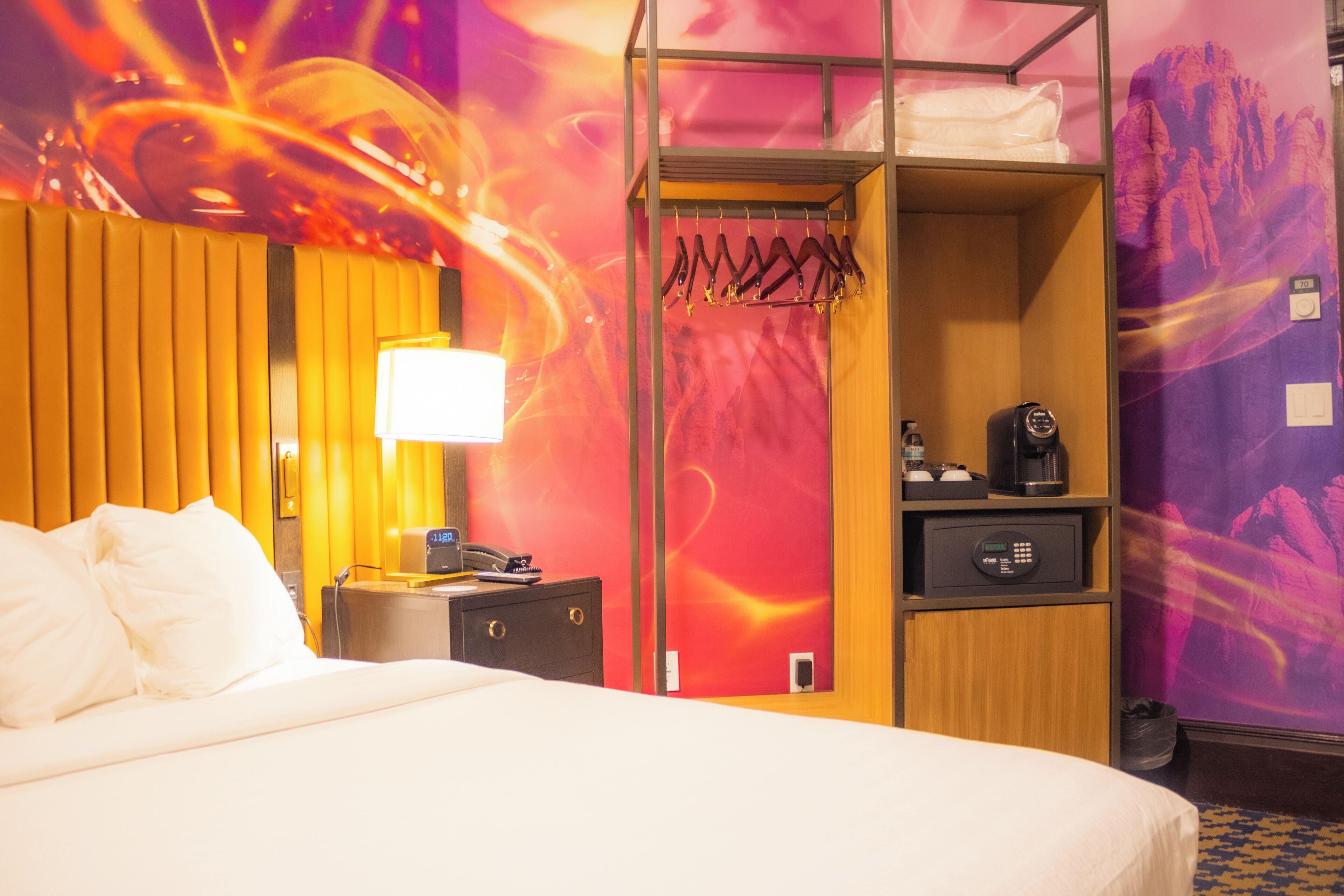 Et hotelværelse med en seng, et natbord, en lampe, et klædeskab og en væg med et farverigt abstrakt design.