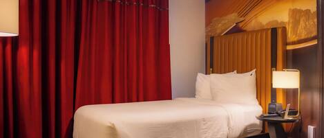 Habitación de hotel con cama, mesita de noche con lámpara y cortina roja.