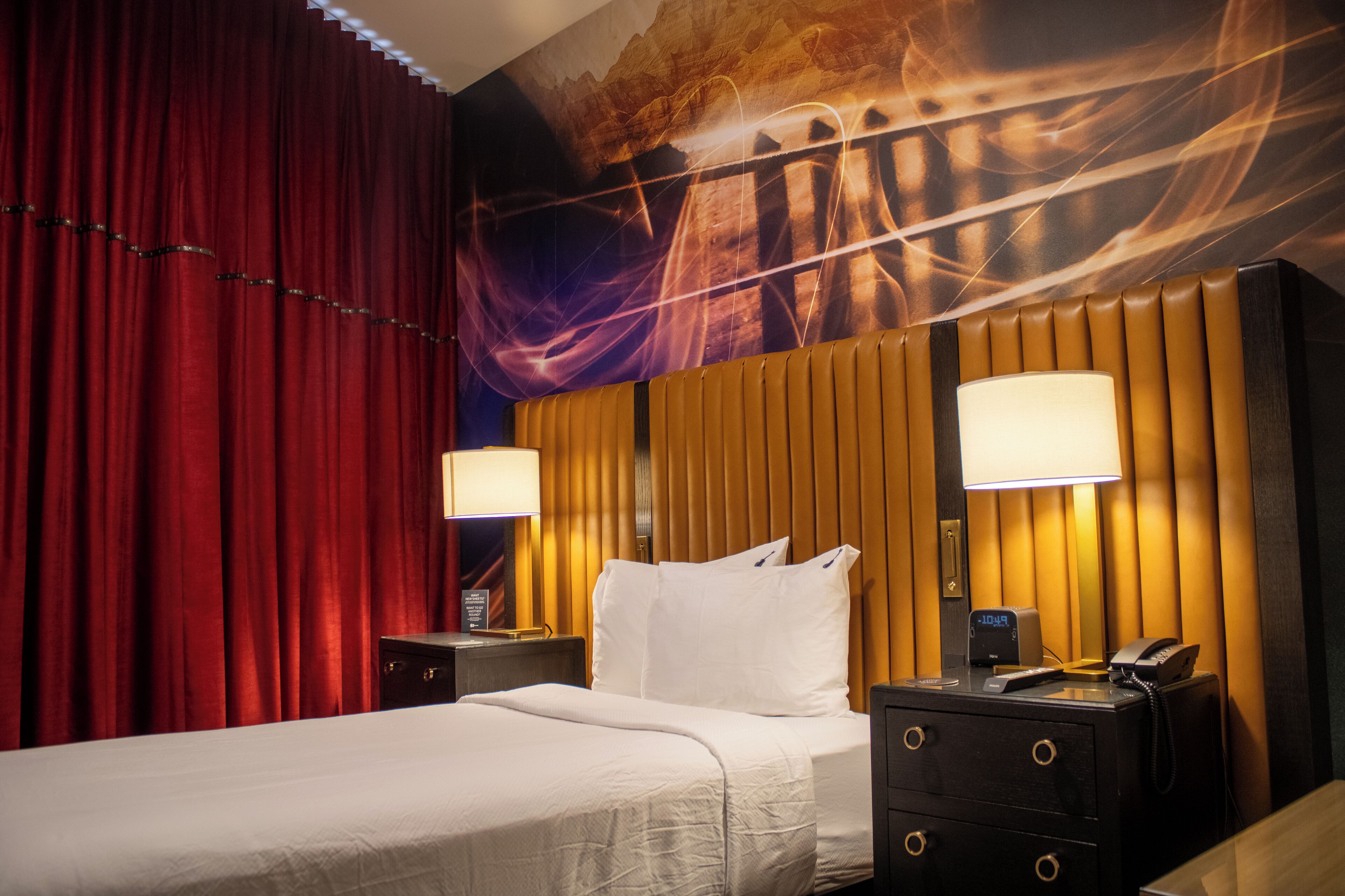 A hotel room with a bed, two bedside tables, a wall-mounted lamp, a telephone, and a red curtain.