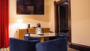 Et hotelværelse med et rundt glastbord, blå polstrede stole, et fladskærms-TV monteret på væggen, et træskrivebord med en lampe og en rød gardin.
