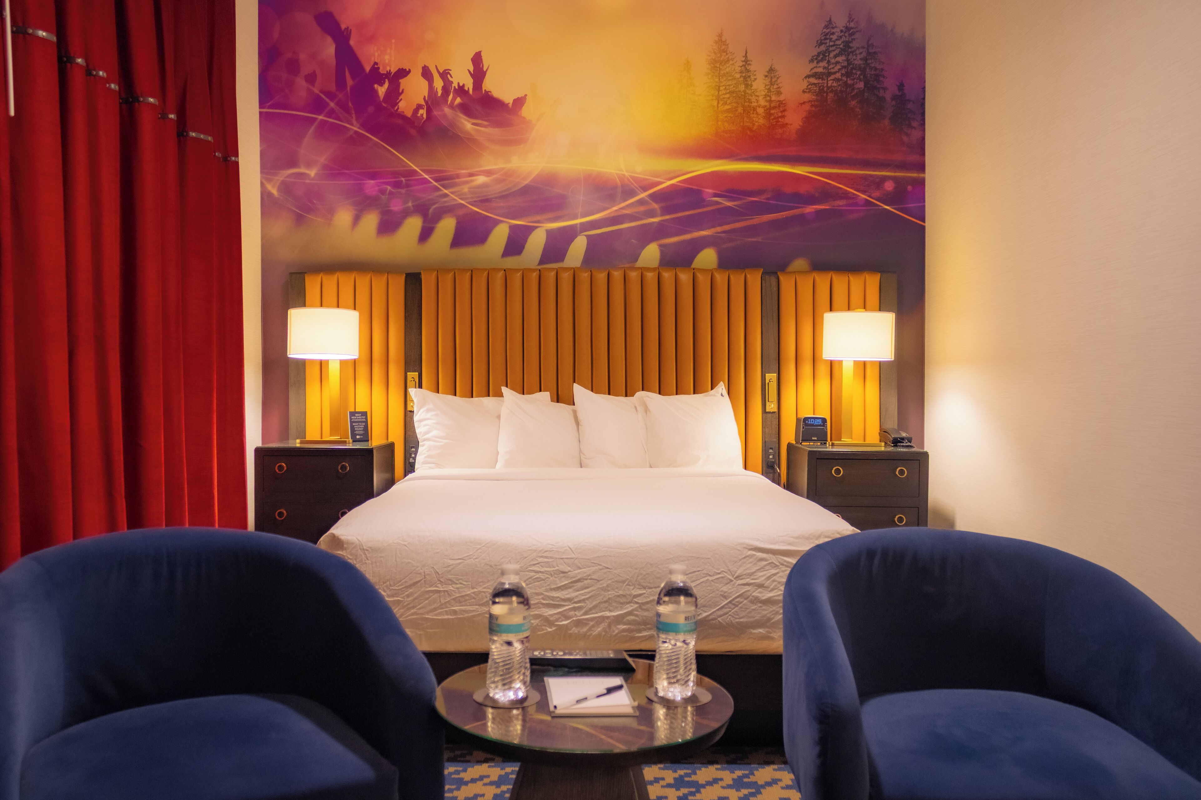 Camera d'albergo con un letto grande, due comodini con lampade, una tenda rossa, due poltrone blu e un affresco.