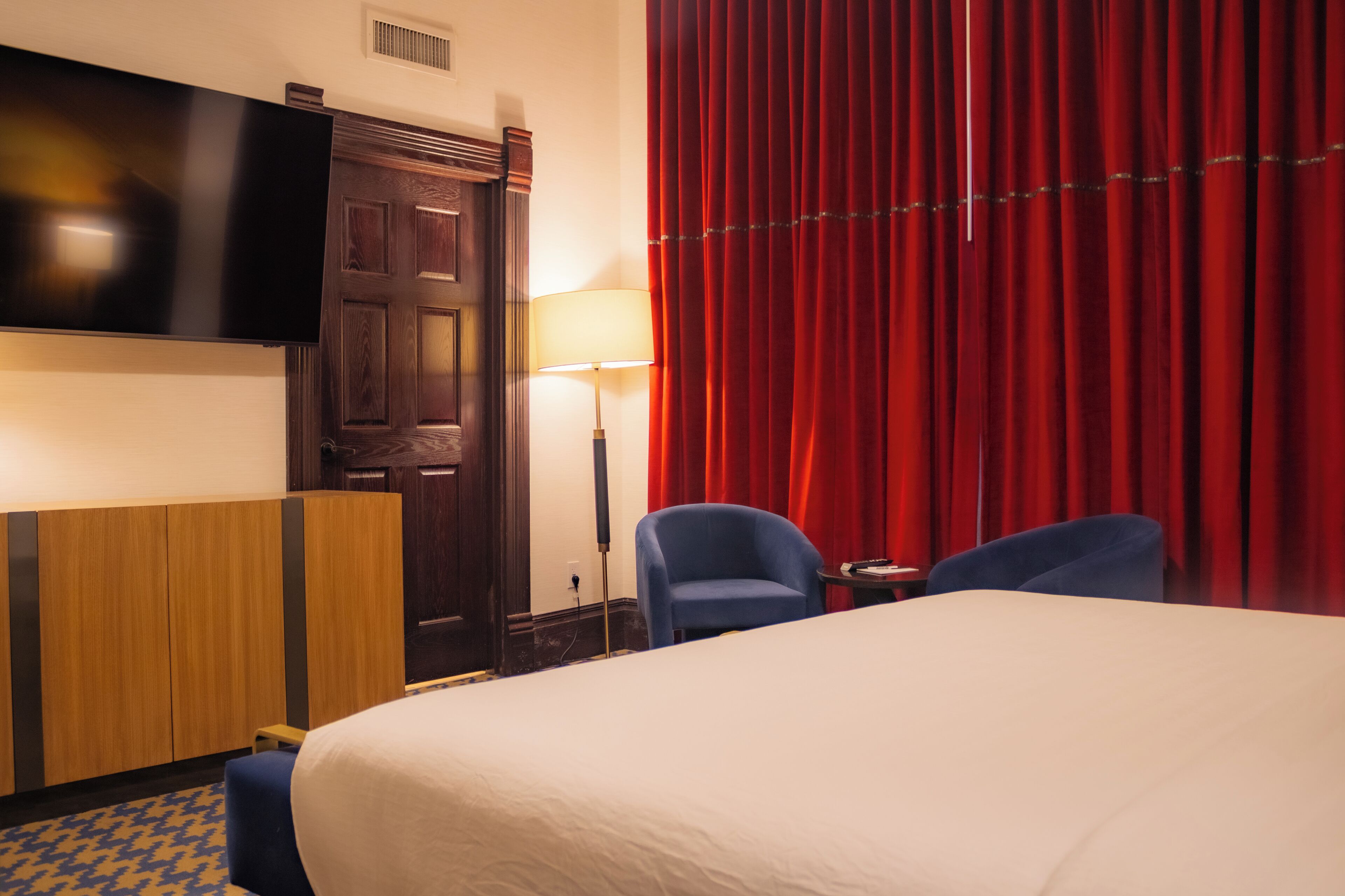 Ein Hotelzimmer mit einem großen Bett, einem flachbildfernseher über einer Holztür, einem Schreibtisch mit Lampe, zwei Stühlen und einem roten Vorhang.