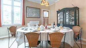 Uma sala de jantar com uma mesa redonda posta para seis pessoas, rodeada por cadeiras de vime. Há um aparador com portas de vidro à direita, e uma obra de arte emoldurada na parede acima da porta.