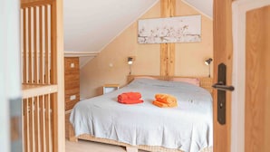 Um quarto acolhedor com teto inclinado, uma cama com manta cinza, duas toalhas enroladas e uma obra de arte fixada na parede.