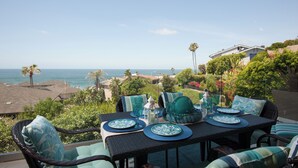 Un comedor con vistas al océano, muebles de exterior y una mesa dispuesta con platos y vasos.
