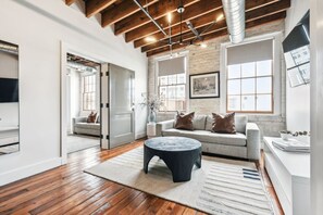 Ein geräumiges Wohnzimmer mit Holzboden, einem runden Couchtisch, einem grauen Sofa mit braunen Kissen und einem großen Fenster, das für natürliches Licht sorgt.
