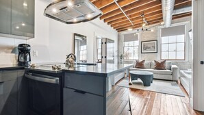 A modern kitchen with a central island, wooden beams on the ceiling, and a large window providing natural light. - Live the Jazz Life: Modern 4BR Condo in NOLA (New Orleans)