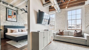 A modern bedroom with a bed, a grey sofa, a TV mounted on the wall, and a large window. - Live the Jazz Life: Modern 4BR Condo in NOLA (New Orleans)