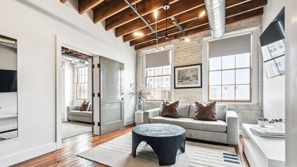 A spacious living room with a wooden floor, a round coffee table, a grey sofa with brown cushions, and a large window providing natural light.