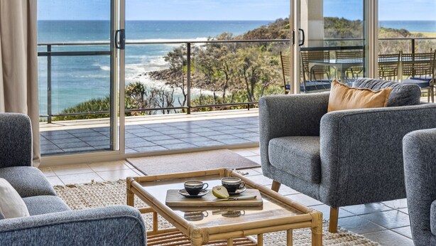 Un moderno salón con un amplio ventanal que da a un balcón con vistas al océano y la playa.