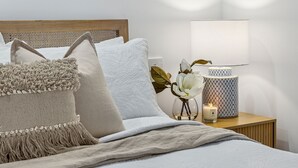 Una cama bien tendida con cabecera de madera, varios almohadones, un plaid suave, una mesita de noche con lámpara, un florero con flores y una vela.