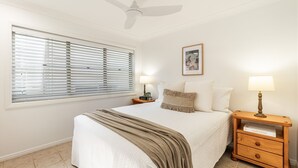 Ein Schlafzimmer mit einem Bett, zwei Nachttischen, einem Deckenventilator, einem Fenster mit Jalousien und einem gerahmten Bild an der Wand.