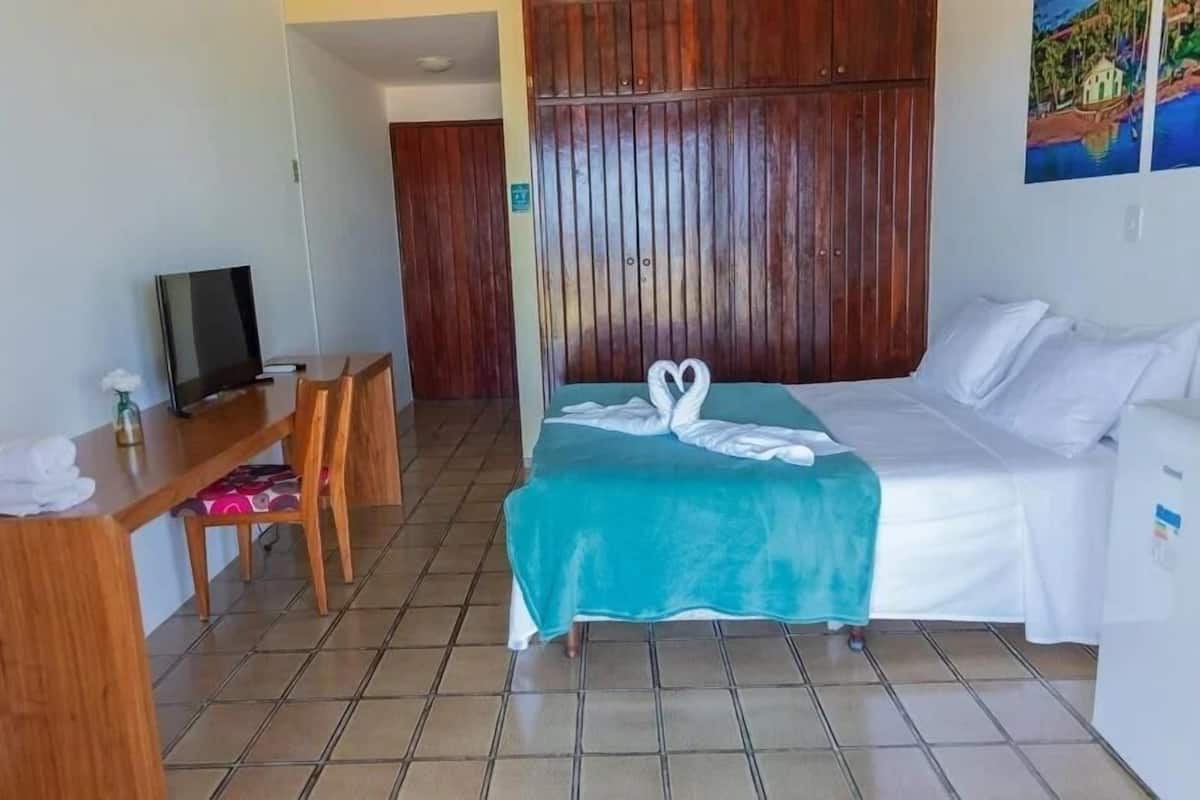 Une chambre d’hôtel avec un lit, un bureau avec une télévision, une armoire en bois et un tableau au mur.