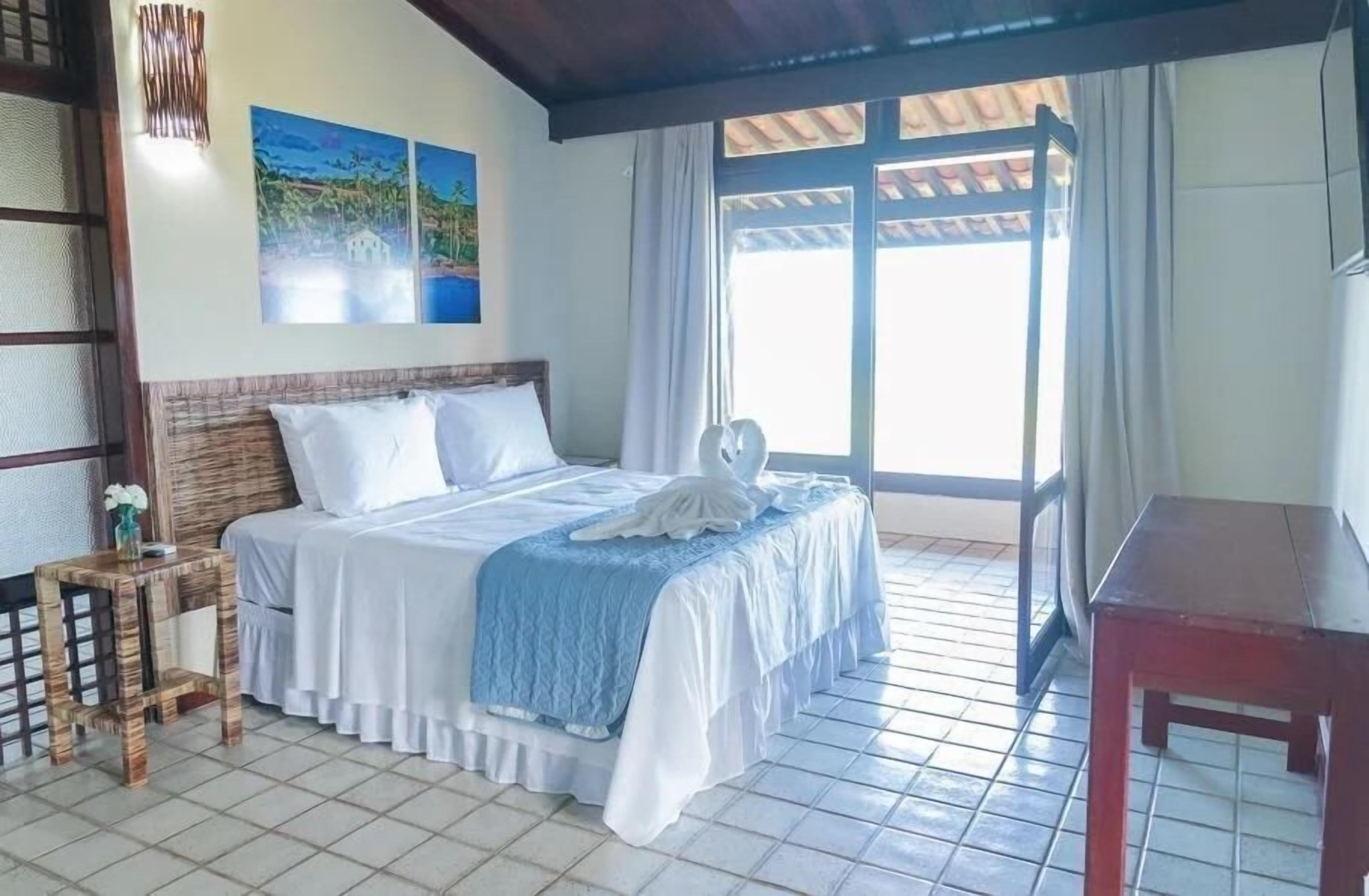 Un dormitorio con cama, mesita de noche, una pequeña mesa y una ventana con cortinas.