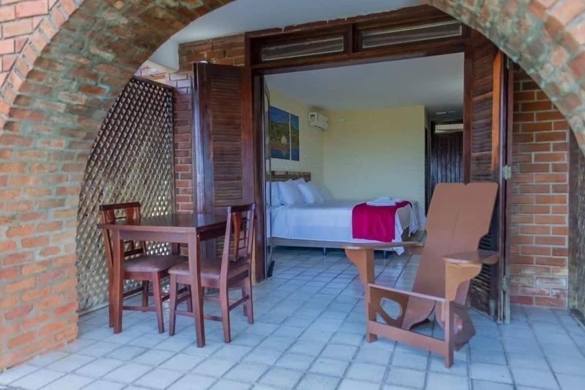 Une terrasse extérieure avec une table et des chaises en bois, un lit avec une couverture rouge et un mur en briques.