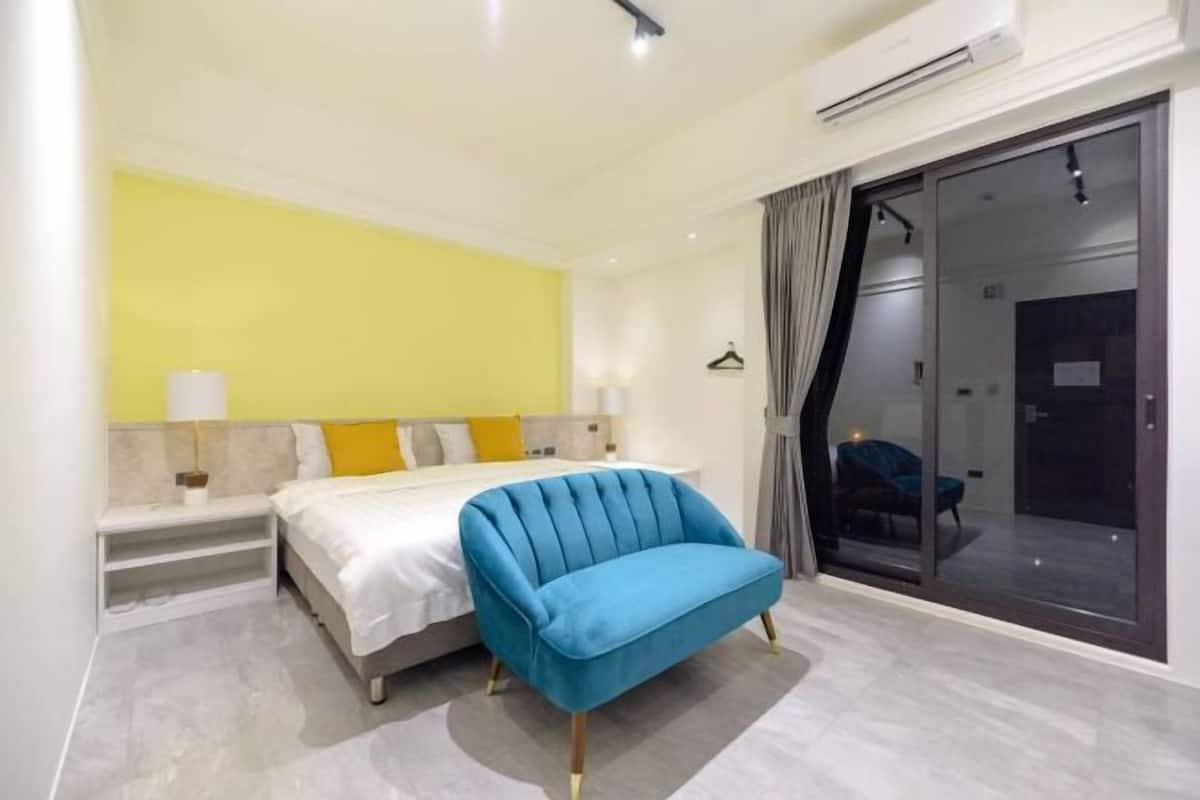 A modern hotel room with a bed, a blue armchair, a bedside table, a lamp, and a sliding glass door leading to a balcony.