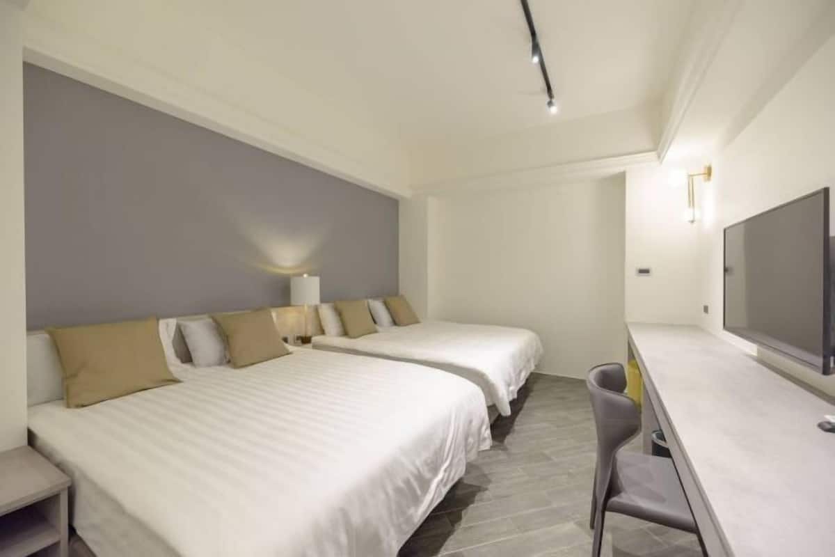 A hotel room with two beds, a desk, a chair, a TV, and a lamp.