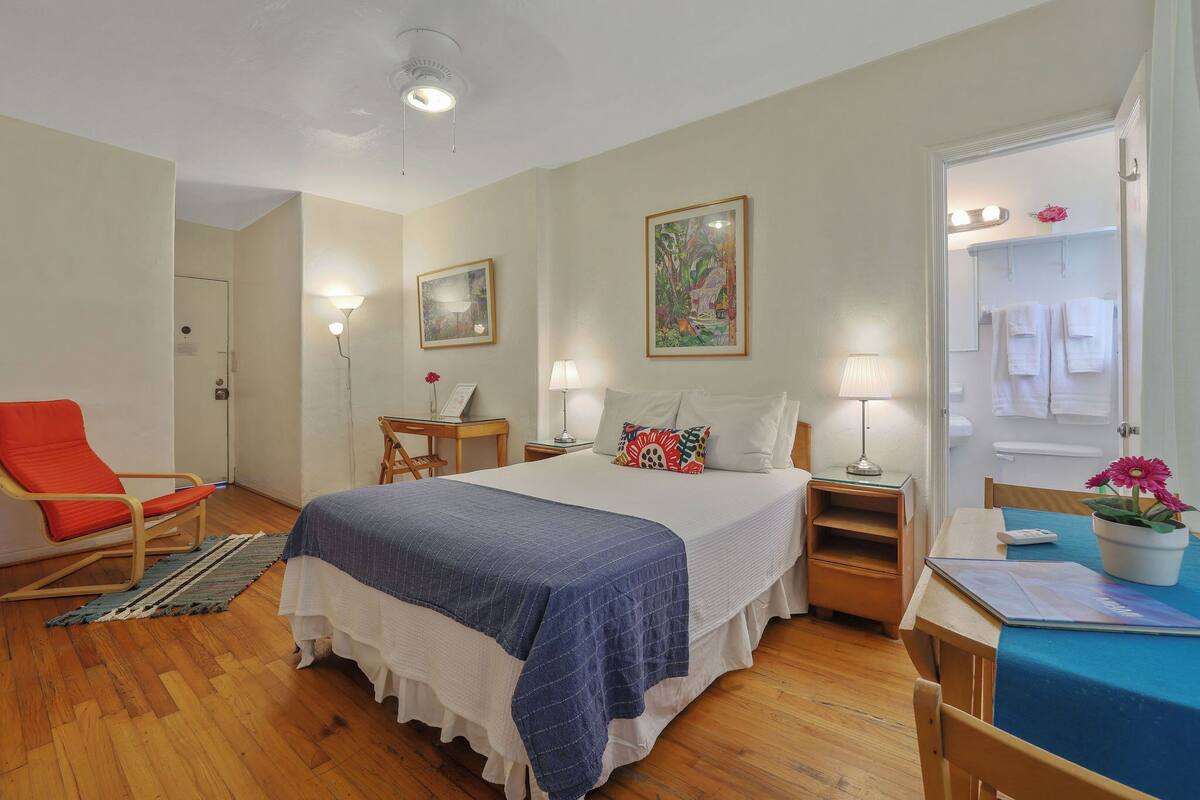 Een slaapkamer met een bed, een stoel, een bureau en een zichtbare badkamer door een open deur.