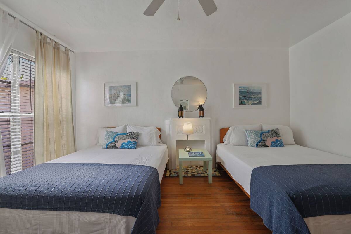Een hotelkamer met twee bedden, een tafeltje met een lamp, een spiegel en twee foto’s in lijsten aan de muur.