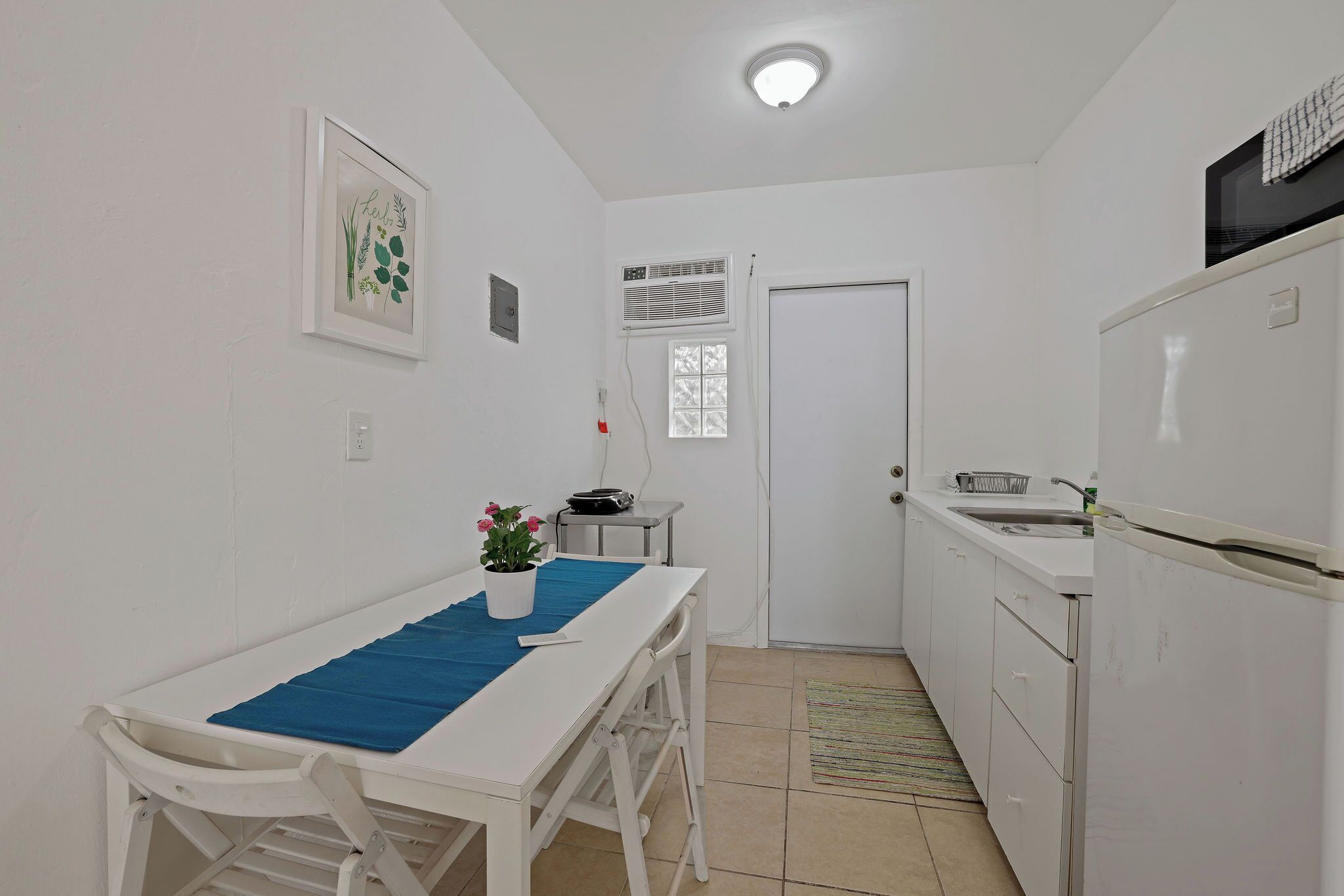 A small, clean kitchen with white cabinets, a white refrigerator, a sink, and a window with blinds. There is a small table with a blue mat and a potted plant.