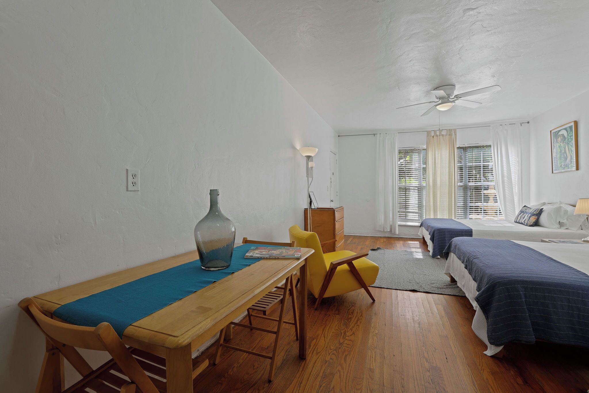A room with a wooden table, a yellow chair, a bed with blue bedding, a ceiling fan, and a window with curtains.