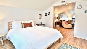 Une chambre avec un grand lit, une petite table de chevet et une chaise. Une porte donne accès à une autre pièce.