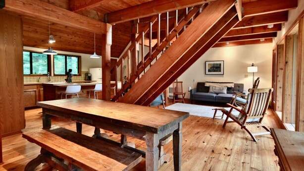 Interior rústico de una cabaña con una mesa de comedor de madera, una escalera y una sala de estar con sofá y sillones.