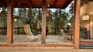 Una terraza de madera con dos sillas y una mesa, rodeada de paredes de vidrio con vistas a árboles.