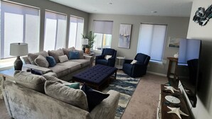 A spacious living room with a large sectional sofa, a blue ottoman, and a coffee table. There are two armchairs and a small side table with a lamp. The room has large windows with blinds, a rug, and a flat-screen TV mounted on the wall. - L`escapade -The Getaway (Lincoln City)