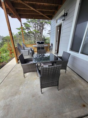 An outdoor patio with a glass-top table and wicker chairs, a grill, and a view of trees and a house. - L`escapade -The Getaway (Lincoln City)