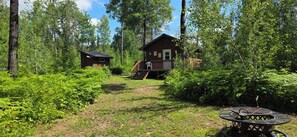 Una cabaña rodeada de un bosque frondoso, con una terraza visible y un cobertizo cercano.