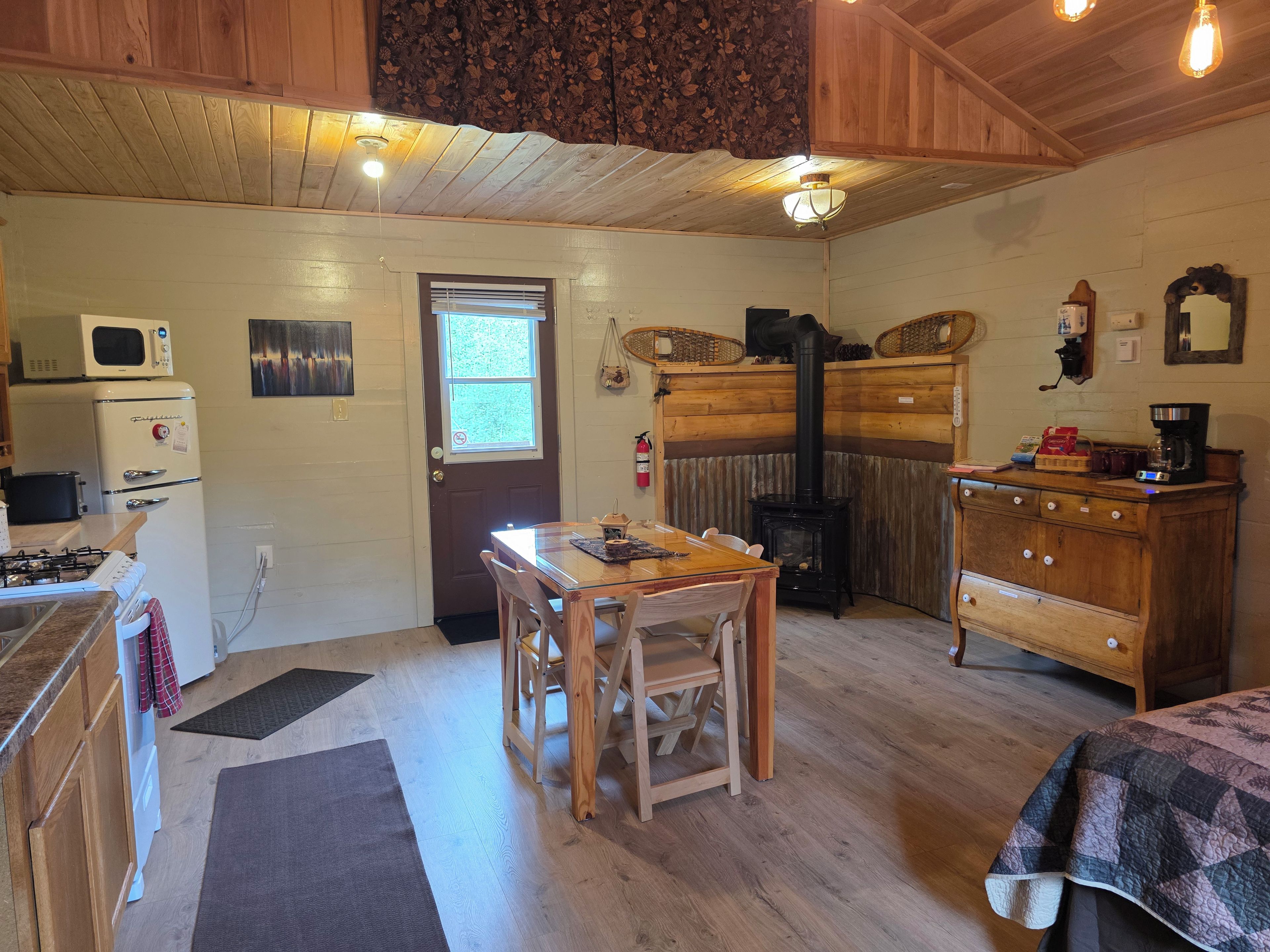 A cozy cabin interior with a dining table, chairs, a wood stove, a kitchen area with a microwave, and a dresser.