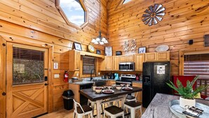 A wooden cabin kitchen with a large window, wooden cabinets, a central island with stools, a microwave, a refrigerator, and a fire extinguisher. - Incredible Vibes, Cozy Fires, and Pure Relaxation! (Sevierville)