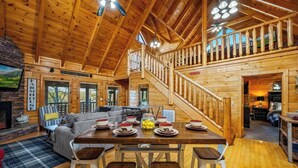 A rustic wooden cabin interior with a dining area, a fireplace, and a staircase leading to an upper level. - Incredible Vibes, Cozy Fires, and Pure Relaxation! (Sevierville)