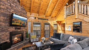 A cozy log cabin interior with a fireplace, a television mounted on a stone wall, and a comfortable seating area with a couch and chairs. - Incredible Vibes, Cozy Fires, and Pure Relaxation! (Sevierville)
