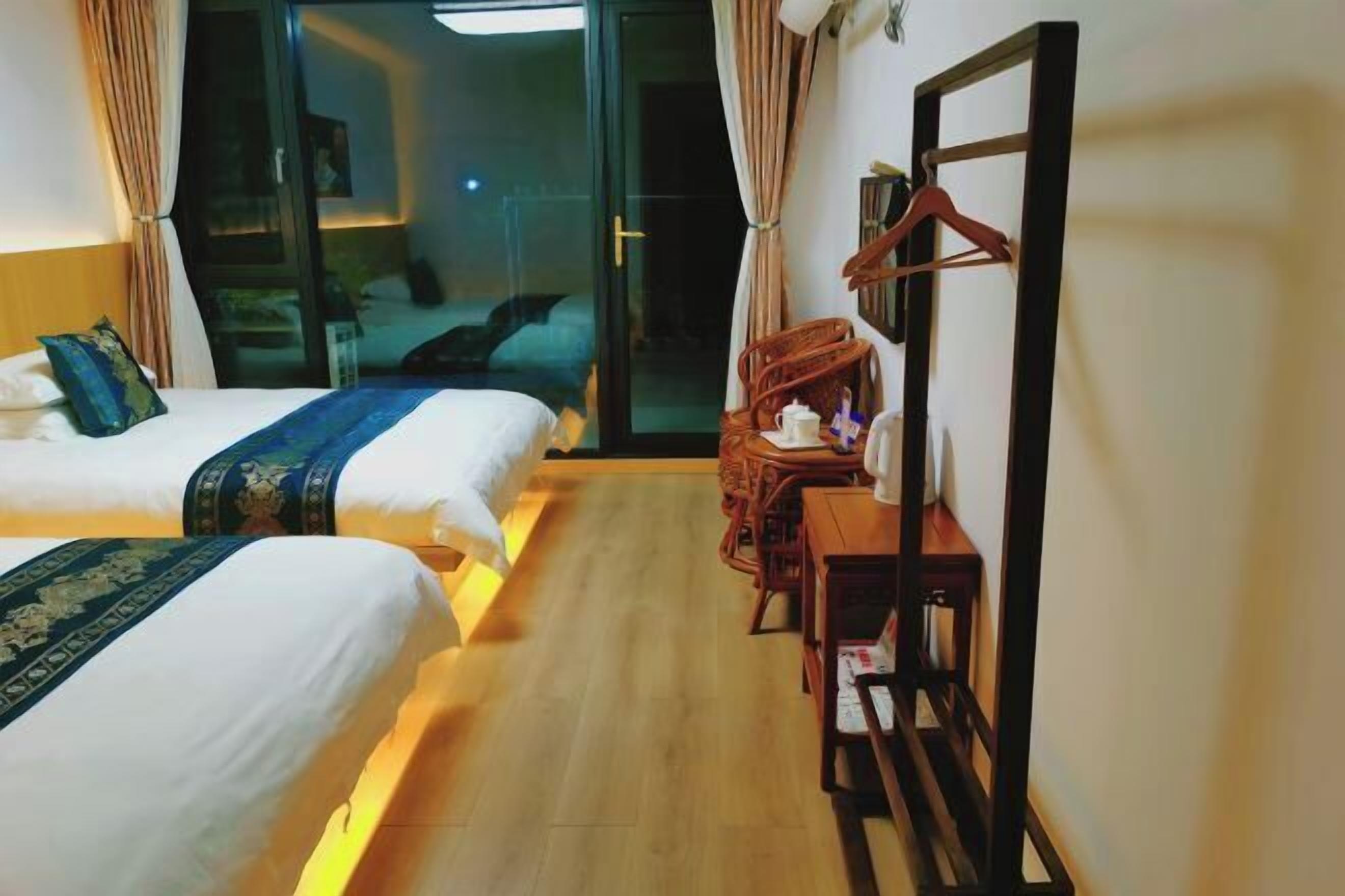 A hotel room with two beds, a wooden floor, a desk with a chair, and a sliding glass door leading to a balcony.