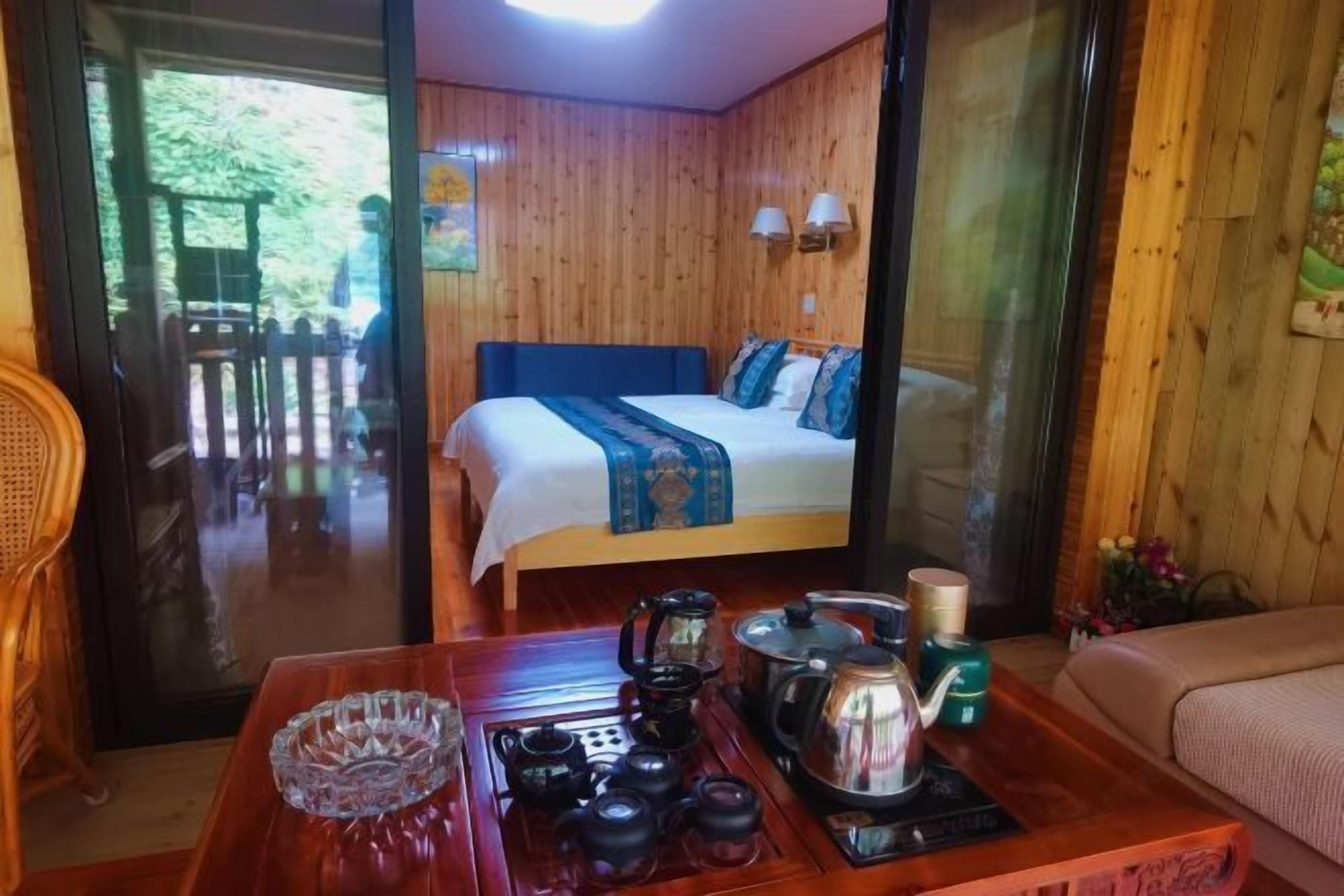 A hotel room with a bed, a wooden table with tea set, and a view of the outdoors.