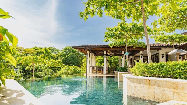 A clear swimming pool with a pergola structure, surrounded by lush greenery and a building with a tiled roof. - Casa Luna at Las Catalinas (Las Catalinas)