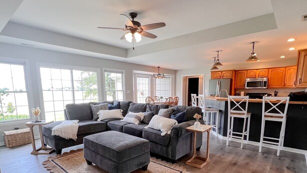 A spacious living room with a large grey sectional sofa, a ceiling fan, and a kitchen area with wooden cabinets and a microwave. - Peaceful - View - Close to Owensboro - Modern Farmhouse (Utica)