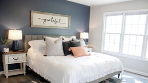 A bedroom with a large bed, two bedside tables, a nightstand, a framed word art, and a window. - Peaceful - View - Close to Owensboro - Modern Farmhouse (Utica)