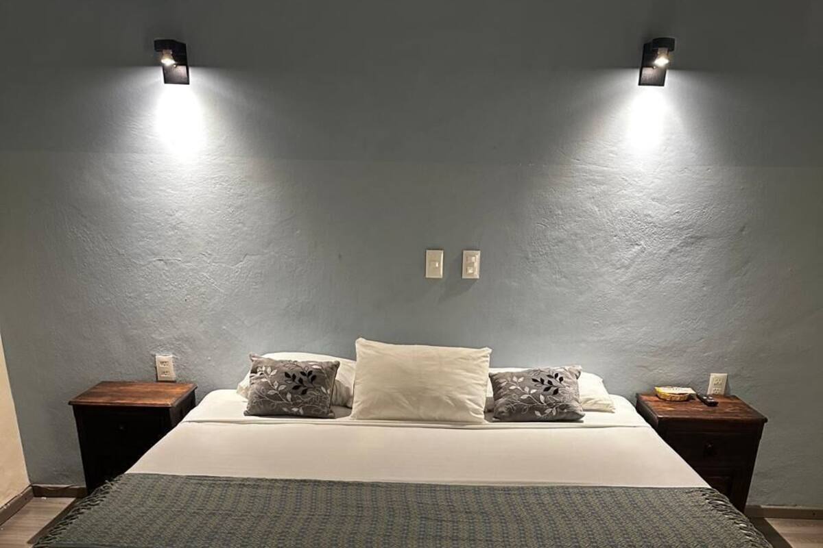 Una cama con colchón blanco y manta gris, flanqueada por dos mesitas de noche con lámparas, frente a una pared gris.