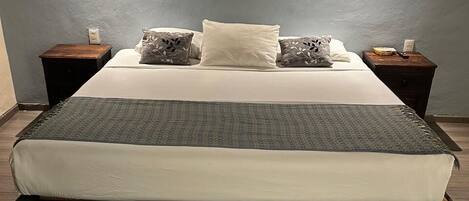 A bed with a white mattress and a grey blanket, flanked by two nightstands with lamps, against a grey wall.